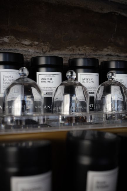 Close-up image of several scented candle jars, some with black lids and others with glass covers, displayed on a reflective surface. The candles are labeled with various names such as 'Smoky Lapasang Sou', 'Oriental Moments', and 'Majestic Earl Grey', indicating different fragrances. The background features a dark, textured brick wall, suggesting an indoor setting. The scene captures the details of the packaging and labels, with soft lighting creating subtle reflections on the glass surfaces. There are no visible furniture or moving activities in this image, but the arrangement of candles is typical of home decor or lifestyle displays, which may be relevant in the context of home moves, packing, or interior reorganisation where decorative items are involved. This image might be used to illustrate the careful packing or handling of personal belongings during a house relocation carried out by professional removals services such as Earls Court Man and Van.