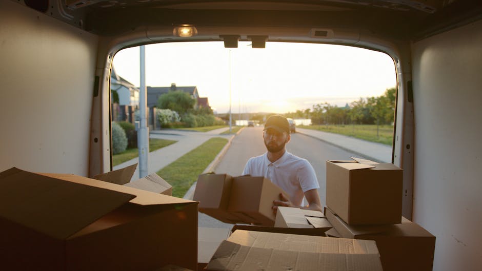 A man wearing a light-colored polo shirt and sunglasses is seen outside the rear of a moving van during sunset, with the van door open, revealing the interior filled with packed cardboard boxes of various sizes. Some boxes are stacked on top of each other, while others are laid out flat or partially opened, indicating they are ready for loading or unloading. The scene is set on a residential street with houses, greenery, and a lamppost visible in the background, and the warm sunlight creates a soft, natural glow. This image captures the process of packing or unpacking during a home relocation, reflecting the services provided by Earls Court Man and Van within their removals operations, including furniture transport and moving logistics.