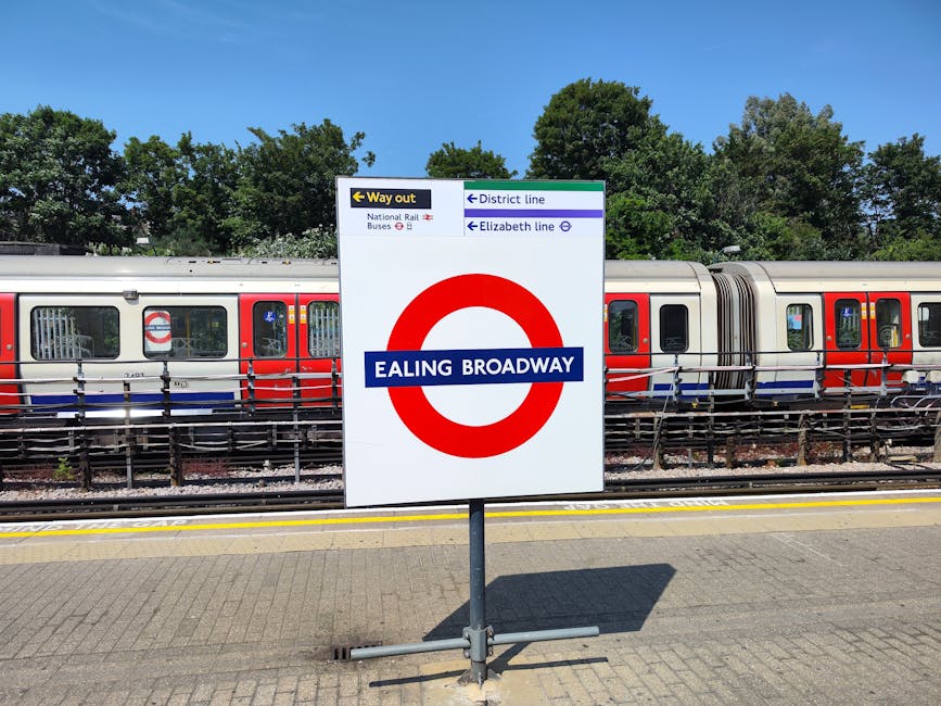 A white sign with a red circle and blue bar displaying 'Ealing Broadway' in white text, positioned on a sidewalk at Ealing Broadway station. Behind the sign, there is a modern London Underground train with red, silver, and white exterior parked on the tracks, with visible windows and doors. To the left of the sign, there are directional signs indicating the way out, with labels for 'National Rail Buses,' 'District line,' and 'Elizabeth line,' alongside arrows pointing left and right. The ground is paved with grey bricks, and a yellow safety line runs parallel to the train. In the background, lush green trees are visible under a clear blue sky, suggesting a bright, sunny day. This scene relates to local transportation and station accessibility, relevant to home relocation and logistical planning provided by Earls Court Man and Van for moving services near Earls Court.