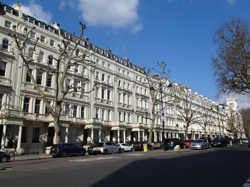 An exterior view of a white Victorian-style terraced building with multiple stories, featuring decorative window frames and balconies. Several leafless trees line the sidewalk in front of the building, with their branches stretching over parked cars, including sedans and hatchbacks, along the street. The sky is partly cloudy with patches of blue visible. The scene depicts a typical residential or mixed-use area in Earls Court, suitable for house removals and furniture transport, and may reflect a location where home relocation services by Earls Court Man and Van are provided. The image captures a calm street setting during daytime with natural lighting, showing the environment where furniture and boxes might be loaded onto a van as part of a professional moving process.