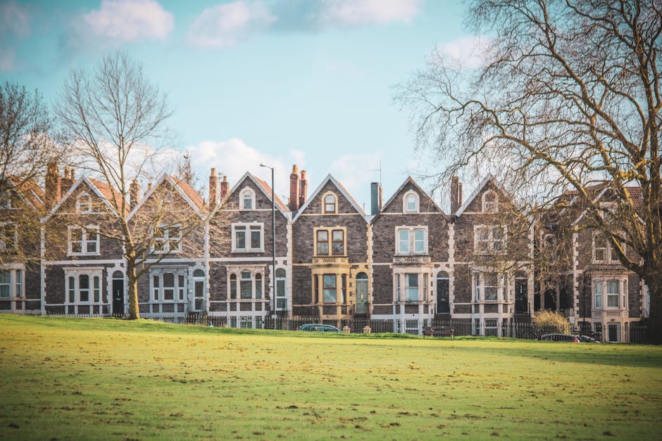 A row of traditional Victorian-style terraced houses constructed from grey stone with white window frames and dark doors, situated behind a green park lawn with a few leafless trees in the foreground. The houses are three to four stories tall with pitched roofs, chimneys, and decorative elements around the windows, reflecting classic residential architecture typical of the Earls Court area. The scene is illuminated by natural daylight under a clear blue sky with some scattered clouds. This setting represents the type of property where professional removals services, such as those provided by Earls Court Man and Van, may assist with home relocations, furniture transport, and packing processes. The peaceful, suburban street environment highlights the context of house move logistics, often involving moving boxes, packing materials, and furniture items to be carefully loaded into a removal van for transportation.