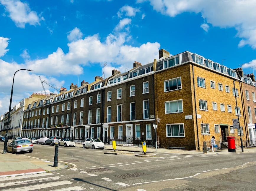 Photograph of a multi-story residential building on West Cromwell Road seen from the street, showcasing a yellow brick exterior with numerous white-framed windows, some with bay windows on the upper floors, and a flat roof with dormer windows. The street scene includes parked cars along the curb, a black lamppost, and a red post box at the corner. Pedestrians are walking on the sidewalk, and a construction crane is visible in the background against a blue sky with scattered clouds. The image captures the urban environment typical of a London neighbourhood, relevant for house removals and client relocation services provided by Earls Court Man and Van, highlighting the context of home moving logistics in this area.