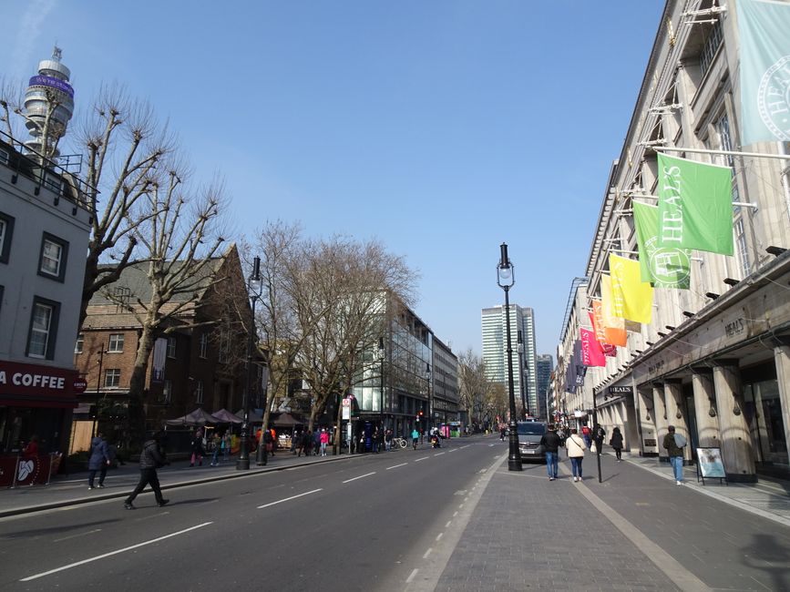 A wide daytime street view of West Cromwell Road in Earls Court, showing a mix of commercial and residential buildings on both sides. On the right, there is a large building with decorative stone facades, multiple colourful banners hanging from the upper floors, and retail stores visible at street level. Pedestrians are walking along the paved pavement, some engaged in conversations or carrying shopping bags. On the left, there are leafless trees lining the sidewalk adjacent to smaller buildings, including a cafe with a red sign. The street is partially occupied by parked cars, with some moving traffic in the distance. The sky above is clear and blue, and a modern high-rise building can be seen further down the street, highlighting the urban environment. The scene captures the bustling atmosphere typical of busy city shopping districts, which can be relevant for illustrating aspects of house removals or furniture transport during a local move in Earls Court, with [COMPANY_NAME] providing professional moving services.
