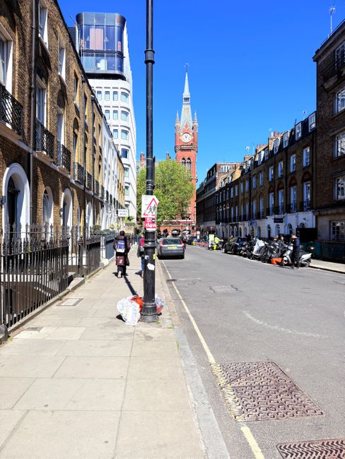 Photograph of a multi-story residential building on West Cromwell Road seen from the street, showcasing a yellow brick exterior with numerous white-framed windows, some with bay windows on the upper floors, and a flat roof with dormer windows. The street scene includes parked cars along the curb, a black lamppost, and a red post box at the corner. Pedestrians are walking on the sidewalk, and a construction crane is visible in the background against a blue sky with scattered clouds. The image captures the urban environment typical of a London neighbourhood, relevant for house removals and client relocation services provided by Earls Court Man and Van, highlighting the context of home moving logistics in this area.